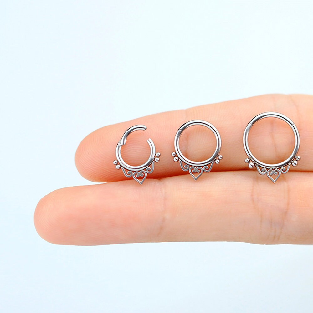 A close-up image of a hand holding three small, circular septum rings from Kaya Movement's "Zura" collection, featuring mandala-inspired designs. The minimalist rings, crafted from 316L steel with a silver look, showcase tiny decorative elements including heart-shaped details at the bottom of each ring. The background is a plain, light blue.